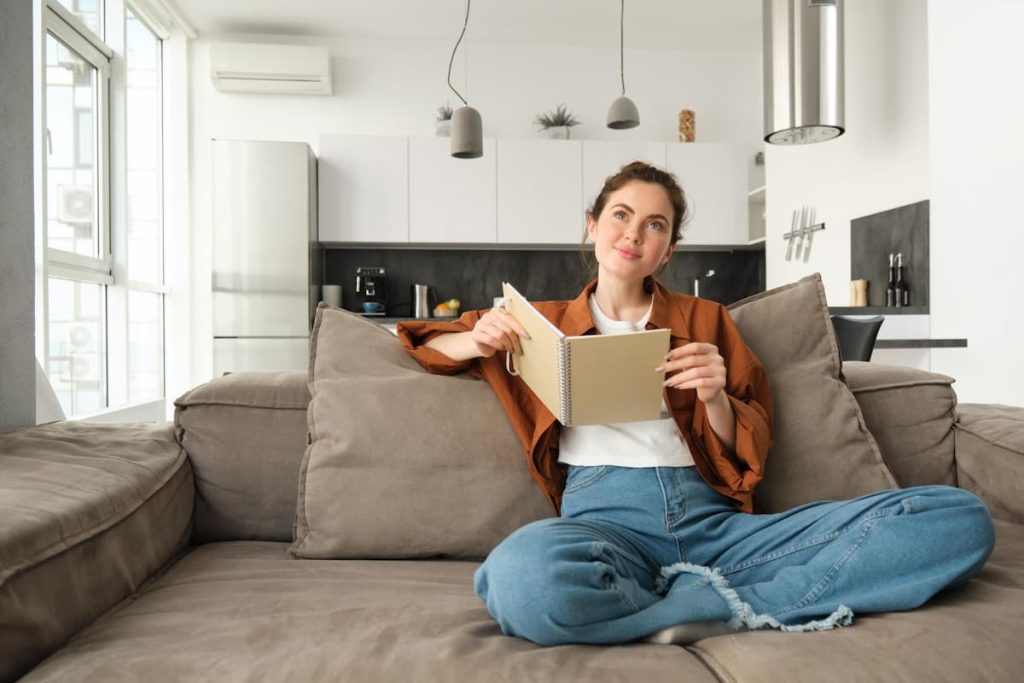 Woman sitting cross-legged on a sofa in a modern living room, holding an open notebook and looking thoughtfully into the distance, perhaps pondering questions about the property or making notes about air conditioning options.