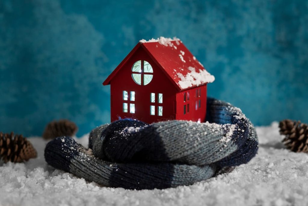 A small red house model surrounded by a blue scarf and artificial snow, with pinecones and a blue textured background, evokes ideas of winter preparation and home comfort improvements.