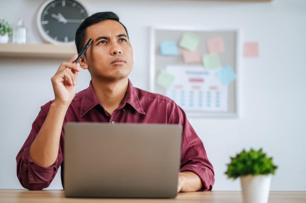 A man sits at a desk with a laptop, holding a pen to his chin and looking thoughtful—perhaps considering ways to improve his business. A clock, calendar, and sticky notes are visible on the wall behind him, hinting at the busy schedule tradesmen often keep while managing tasks like Local SEO.