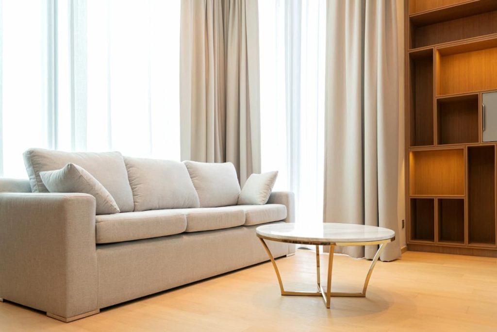 A modern living room featuring a light grey fabric sofa, a round marble coffee table, wooden shelves, and large windows with beige curtains.