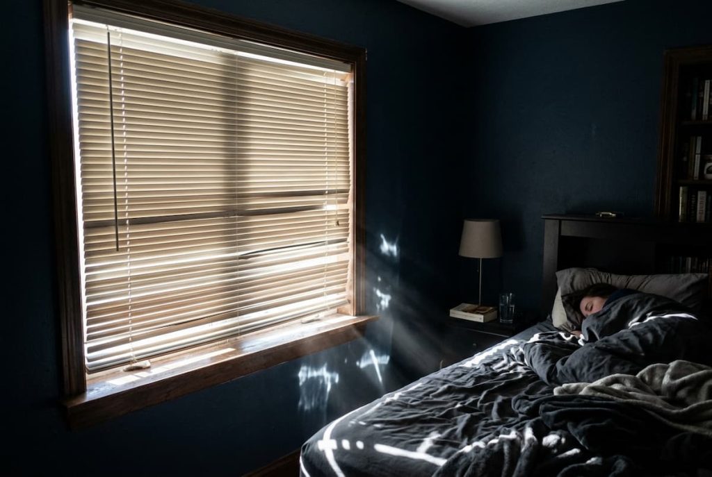 A person sleeps in a dimly lit bedroom, with sunlight streaming through the bedroom blinds, creating light leaks that dance across the bed and walls.