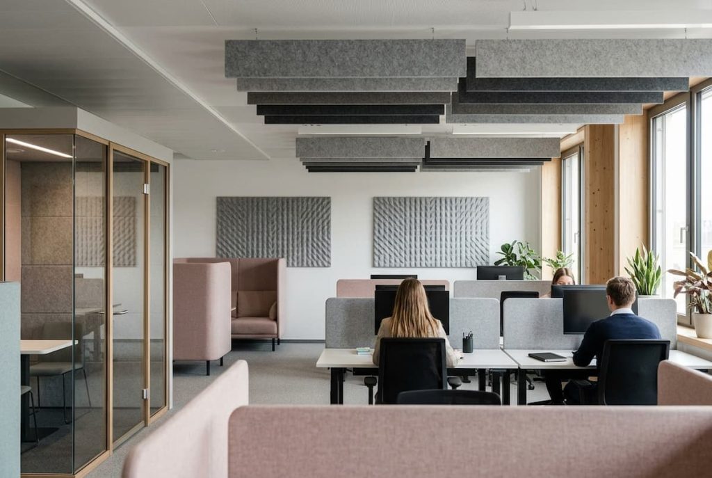 Modern open-plan office with desks, partition panels, acoustic ceiling tiles for improved office acoustics, indoor plants, and several people working at computers. A glass meeting room featuring demountable partitions and a lounge chair are visible on the left.