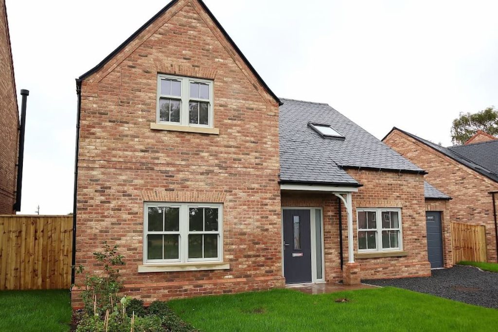 Two-storey brick house with grey roof, white-trimmed heritage-style windows, and a small covered porch, surrounded by a grass lawn and a wooden fence. This charming home blends classic appeal with touches of modern construction.