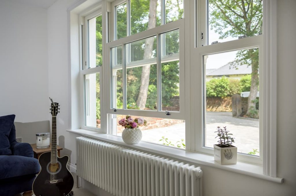 Large white-framed heritage-style windows in a bright room with a radiator below, a vase of flowers, a potted plant, an acoustic guitar, and part of a dark chair visible. Trees and drive outside provide a tranquil view that complements the modern construction within.