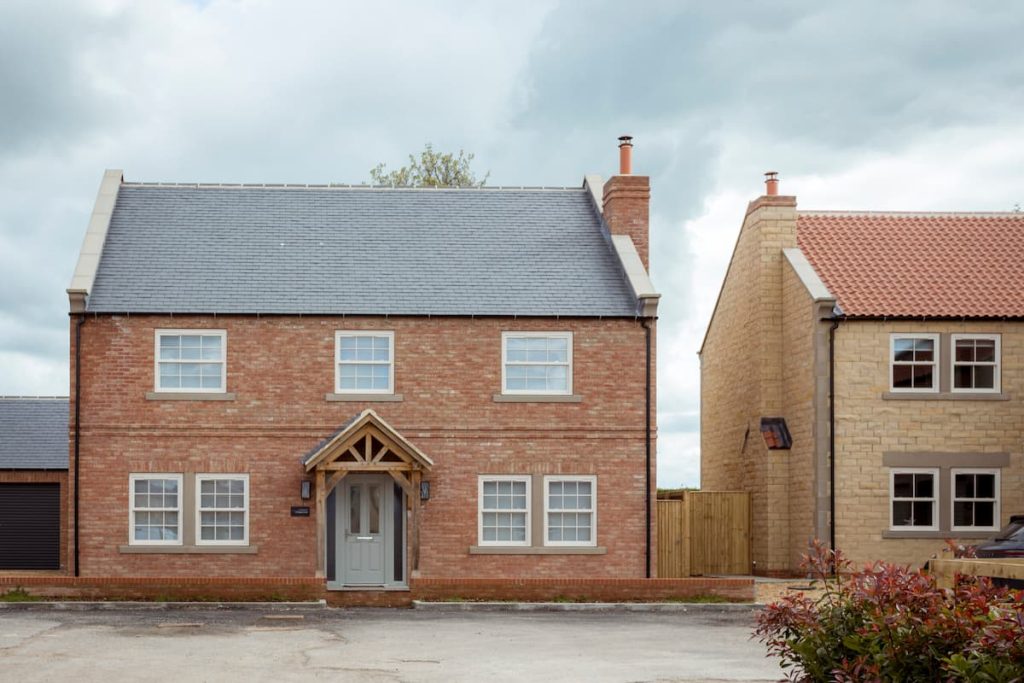 A two-storey brick house with six heritage-style windows and a grey front door, located next to a beige stone house under a cloudy sky. This charming home beautifully blends modern construction with classic window evolution for timeless appeal.