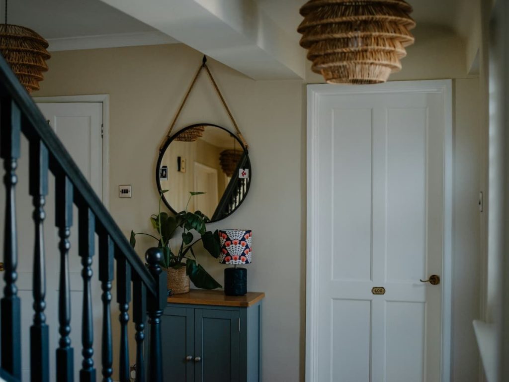 A stylish hallway features a round mirror above a functional blue entrance console table, accompanied by a potted plant, a patterned lamp, two white doors, and basket-style ceiling lights.