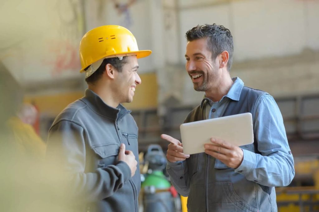 Two men in work uniforms, one wearing a yellow hard hat, smile and talk whilst looking at a tablet in an industrial setting—typical of professional builders or contractors making use of UK construction estimating services.