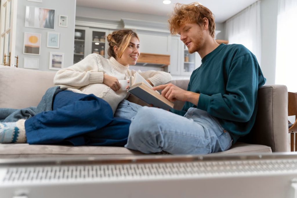 A woman holding a mug and a man holding an open book sit on a sofa, talking and smiling in a bright living room, discussing ideas for their house extension and how to avoid common contractor mistakes.