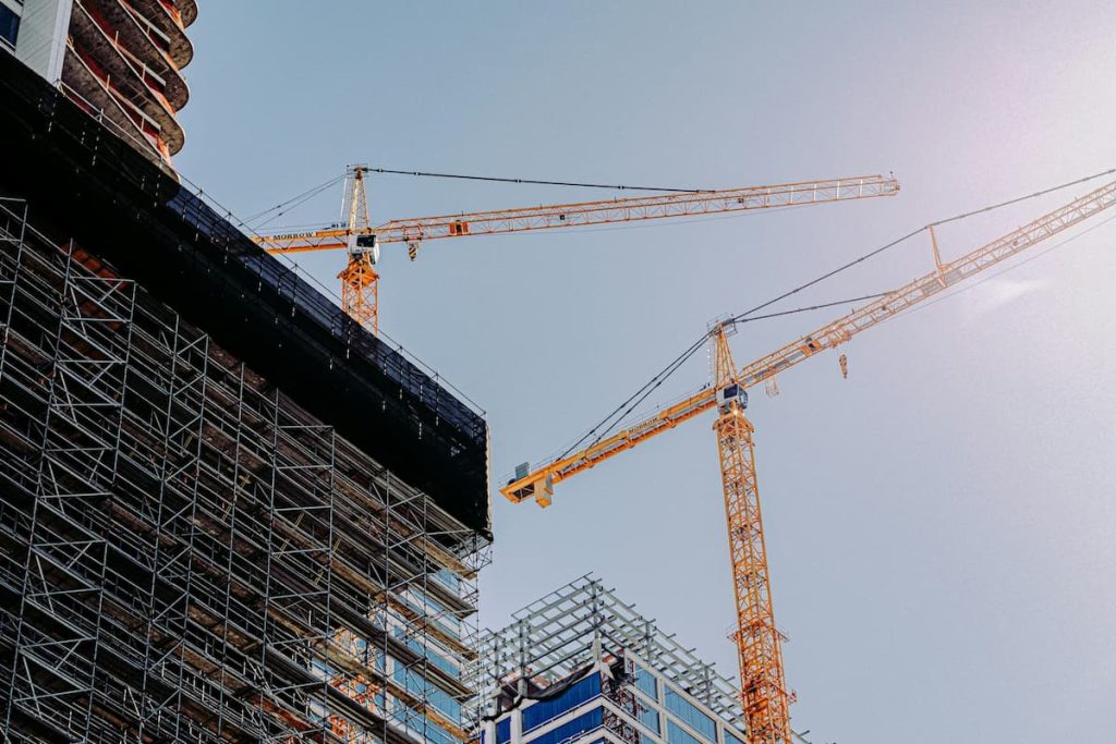 Two yellow construction cranes stand above partially built high-rise buildings with scaffolding, under a clear sky and bright sunlight, showcasing efficient construction scheduling that helps keep builds on schedule.