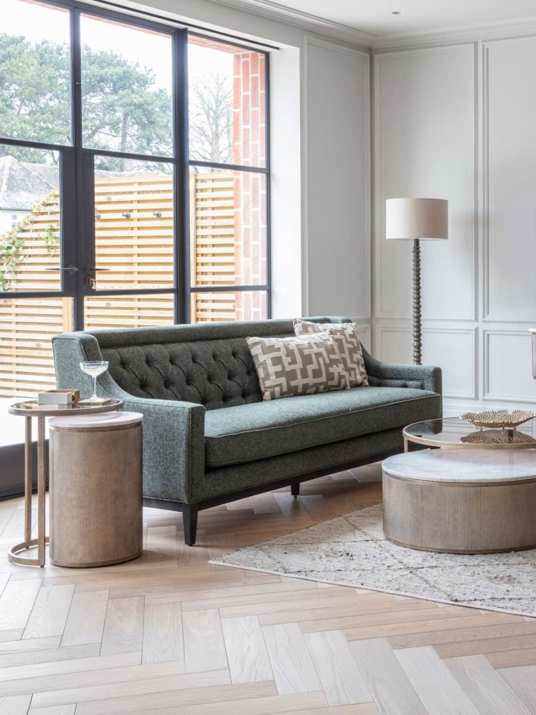 A modern living room featuring a grey tufted fabric sofa, two patterned cushions, a round coffee table, side tables, a floor lamp, and large windows with an outdoor view.