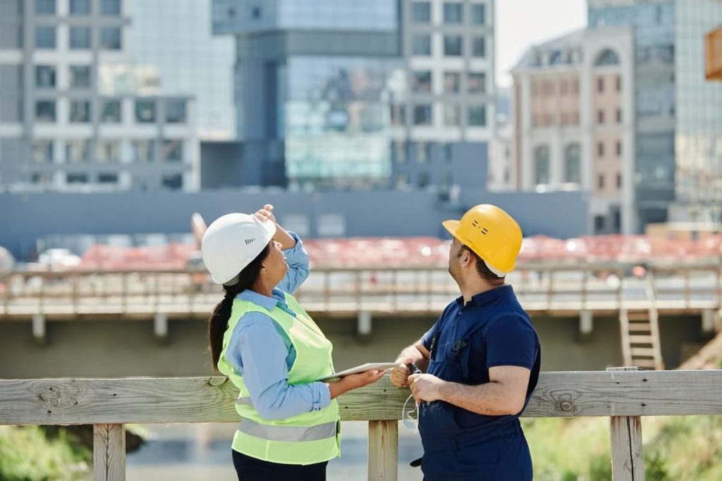 Two construction workers wearing safety helmets and vests stand by a wooden railing, looking and pointing towards buildings in the background at a construction site—a testament to how a thriving construction business can sustain growth whilst shaping the city skyline.