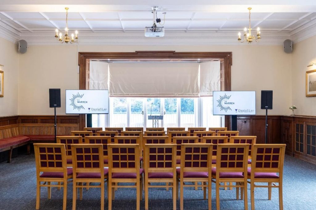 Rows of wooden chairs face two screens and a window in a conference room with wood panelling, chandeliers, and a projector on the ceiling.