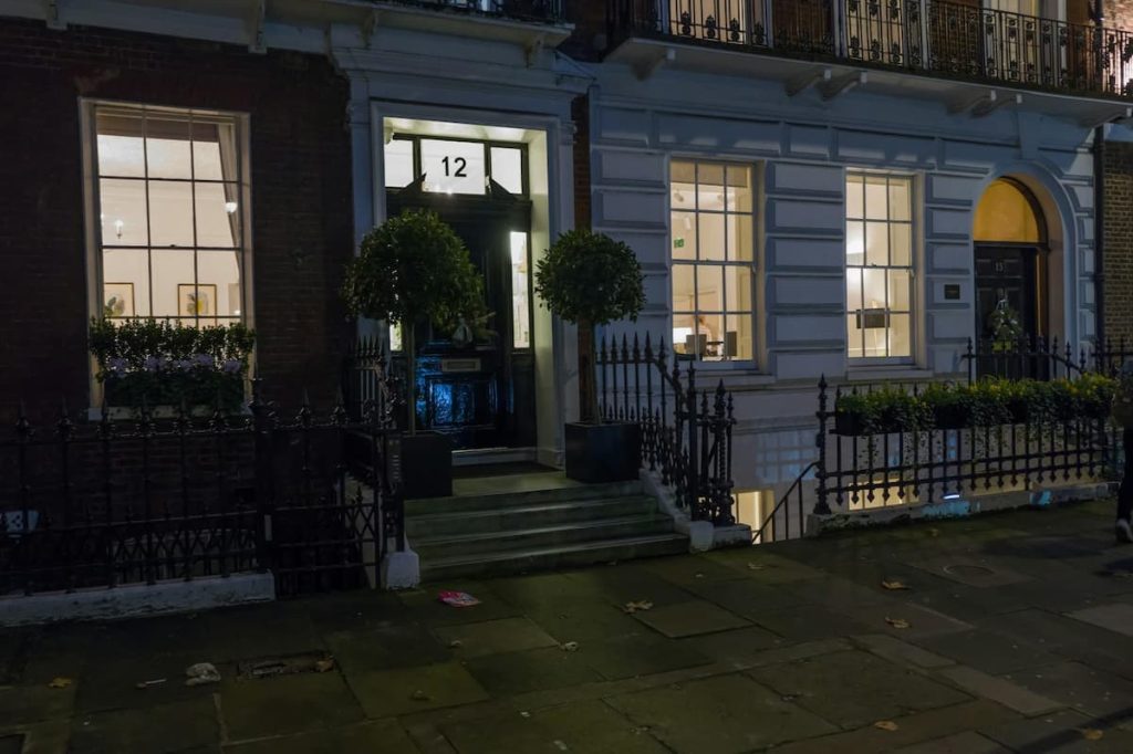 Front entrance of a building at night, with large windows lit from inside, black door labelled “12,” steps, and potted trees on either side of the entrance—showcasing a sophisticated London loft conversion that blends modern design with classic architectural details.
