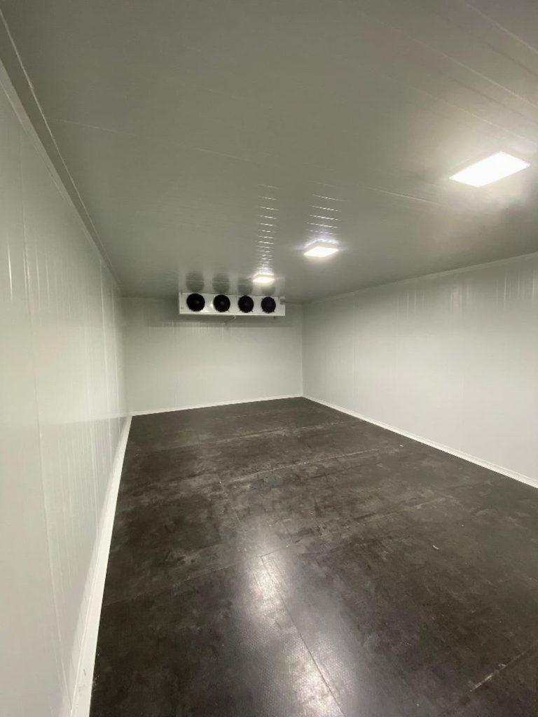 Empty walk-in cold store room with white insulated walls, dark flooring, ceiling lights, and a cooling unit mounted on the back wall—perfect for commercial kitchen cold storage needs or new cold store installation.