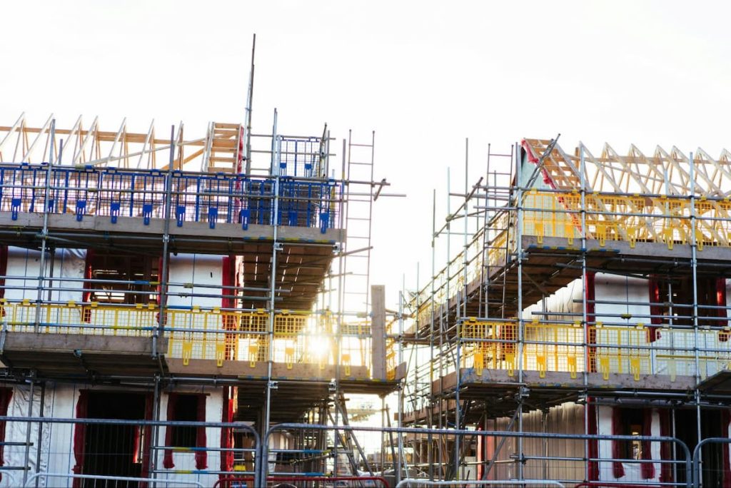 Two partially constructed buildings with scaffolding and exposed wooden roof frames are shown at a building site during daylight, highlighting the £30,000 problem housebuilders face due to Part L rework.