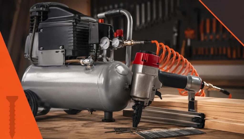 An air compressor with pressure gauges is connected to a pneumatic nail gun by an orange coiled hose, illustrating essential tool requirements for any woodworking project. The pneumatic tools rest on a wooden workbench with nails and wooden planks nearby.