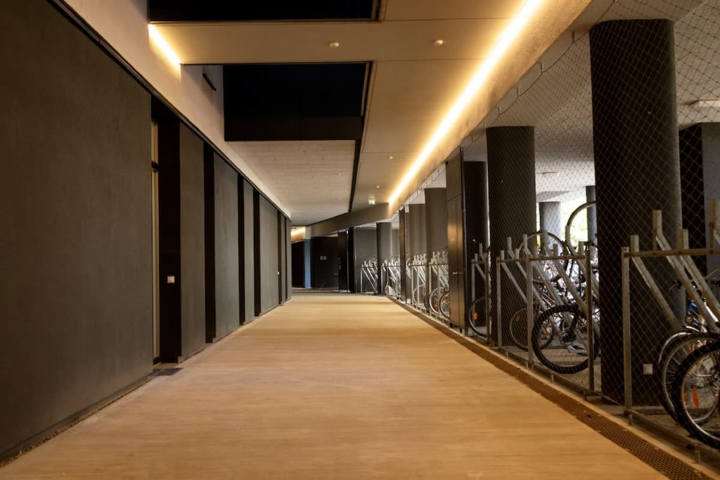 A modern indoor corridor with dark walls and ceiling lights, adjacent to a fenced bicycle parking area with several bikes visible.