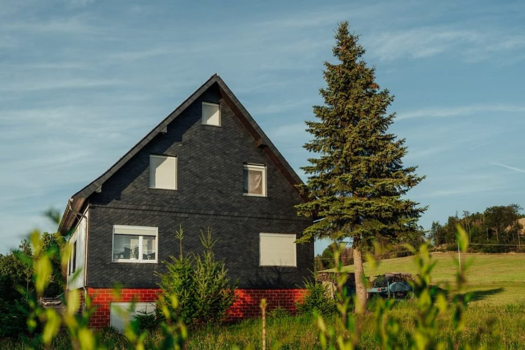 A house built according to Passivhaus principles, with a dark sloping roof and red brick lower walls, stands next to a tall pine tree in a grassy rural area under a blue sky.
