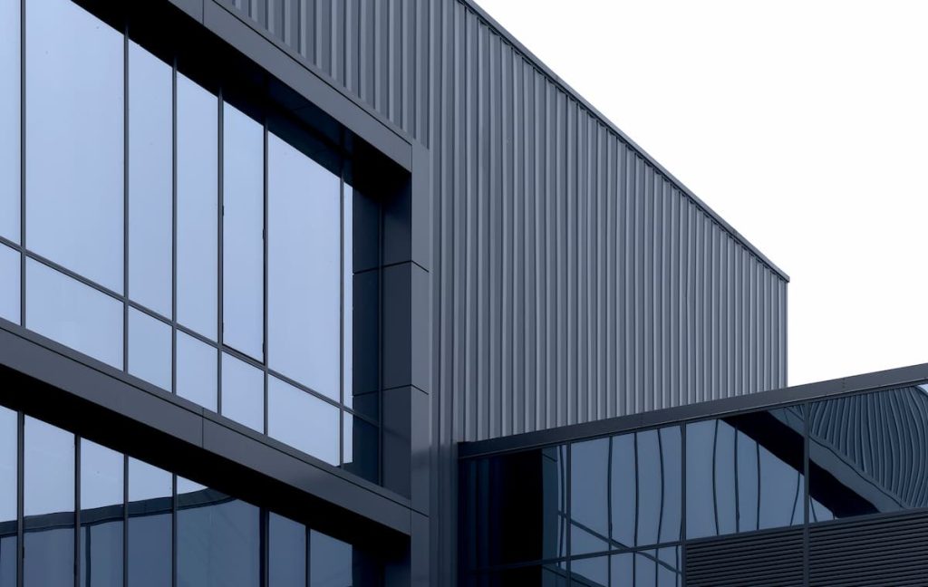 Modern building exterior with large glass windows and aluminium cladding, reflecting the sky; photo taken from a low angle against a white background. The durable cladding material enhances both aesthetics and longevity.