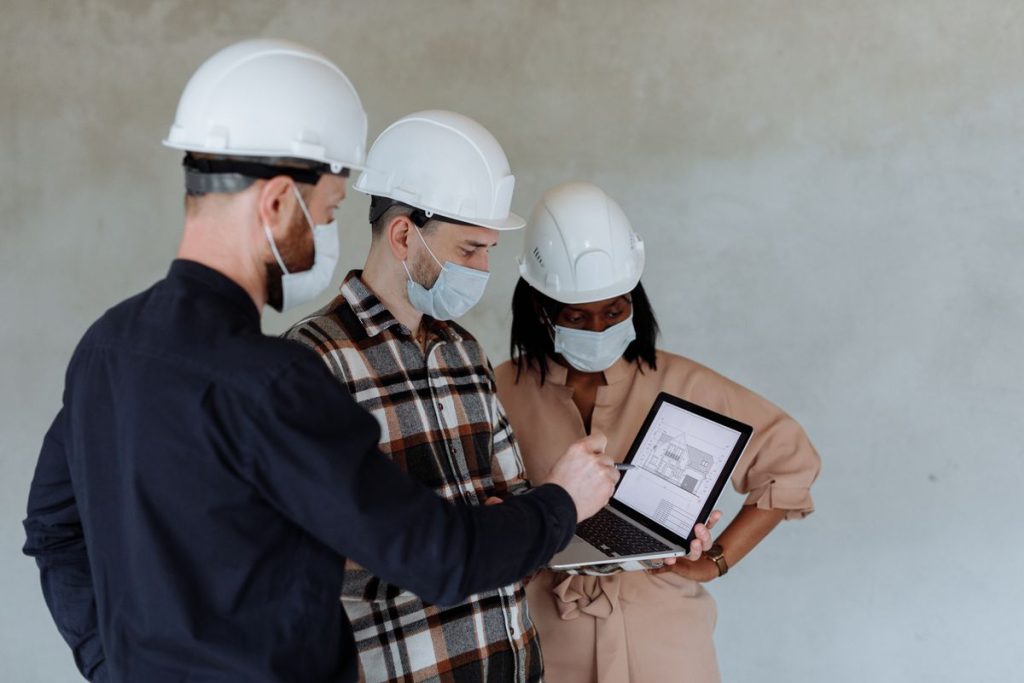 Three people wearing hard hats and masks review architectural plans on a laptop at a construction site, demonstrating how construction companies can streamline operations with an AI-ready sales process.