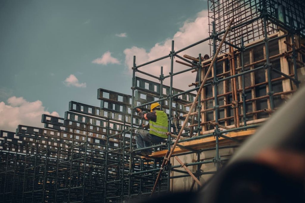 A construction worker in a yellow hard hat and green vest works on scaffolding at a building site under a partly cloudy sky, showcasing the expertise of UK Contractors specialised in projects such as Precast Retaining Walls.