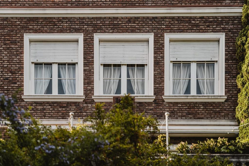 Three sash windows with white shutters and curtains on a dark brick building.