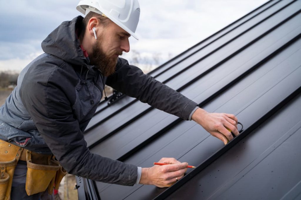A construction worker in a hard hat measures and marks a black metal roof panel with a pencil during a routine roof inspection.