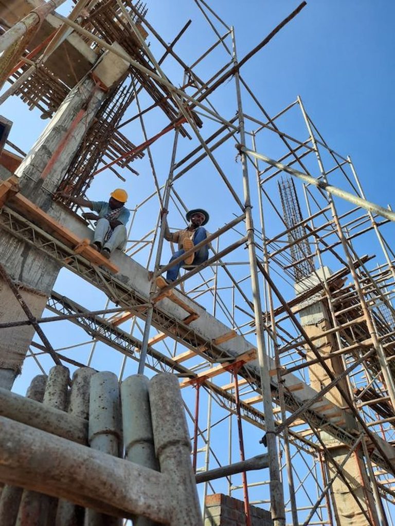 Two construction workers sitting on a concrete beam within a tall metal scaffolding structure, demonstrating site safety ergonomics.
